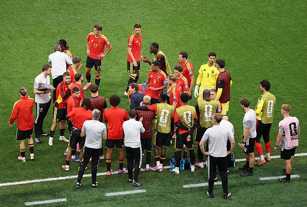 Soccer team huddle on a green field with players in red jerseys listening to coaches and staff in white and gray. Soccer team huddle on a green field with players in red jerseys listening to coaches and staff in white and gray.