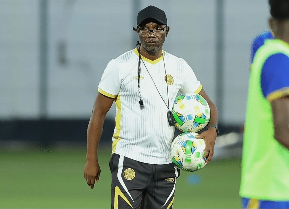 Soccer coach wearing white and yellow kit with whistle, holding two soccer balls on a practice field.