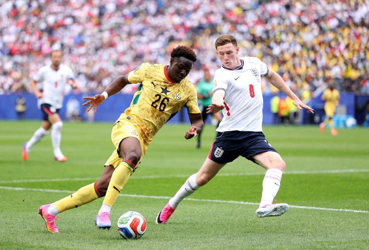 Ghana and England players contesting the ball during a soccer match, with a yellow Ghana kit (number 26) on the left and a white England kit (number 8) on the right on a green field. Ghana and England players contesting the ball during a soccer match, with a yellow Ghana kit (number 26) on the left and a white England kit (number 8) on the right on a green field.