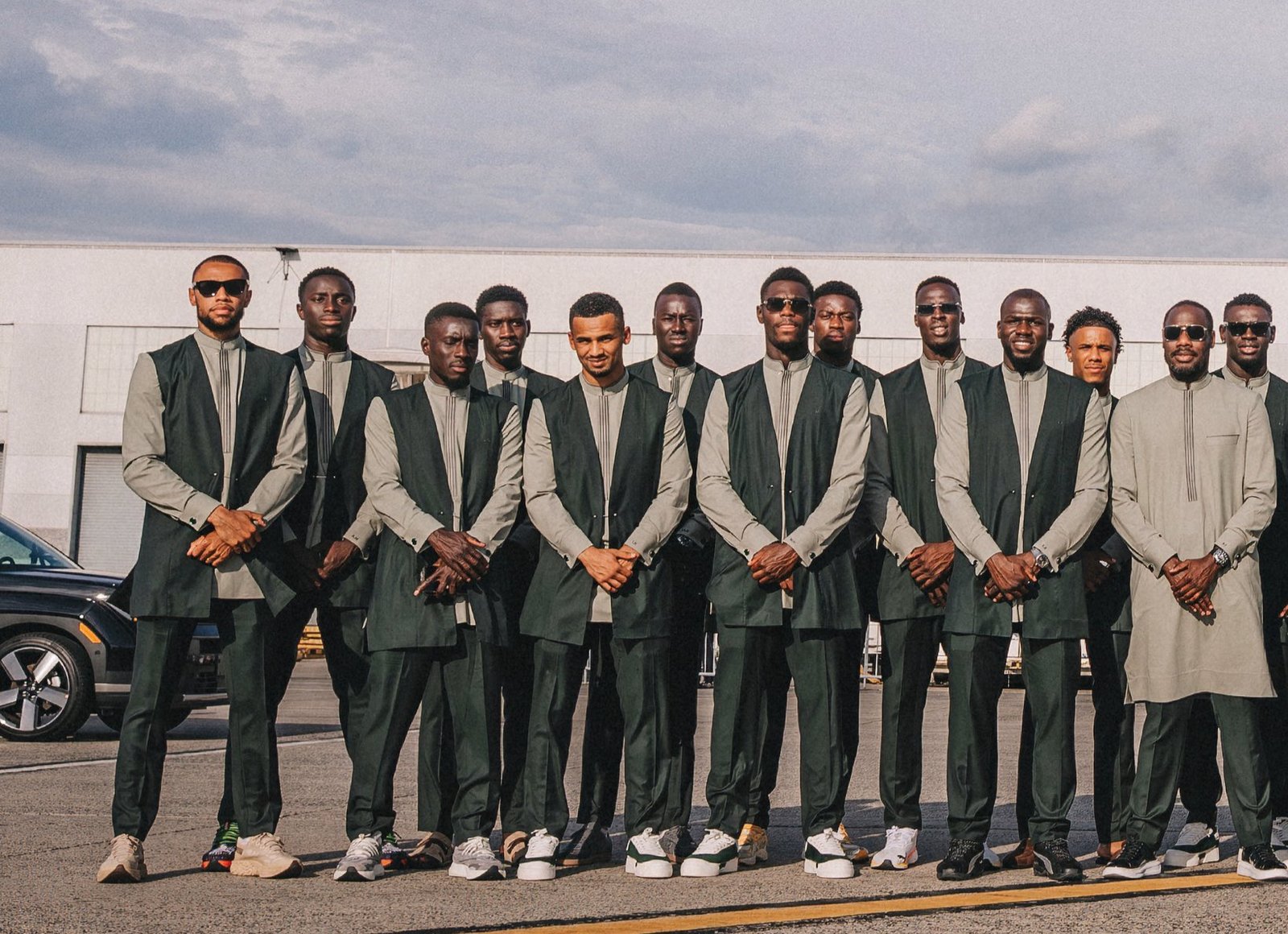 A line of men in matching green and gray outfits poses outside a modern building on a sunny day. sawn, sunglasses on several, hands clasped in front.