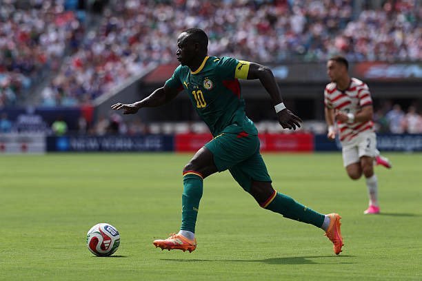 Soccer player in green jersey with number 10 dribbles the ball on a grass field during a match, crowd in stands behind him.