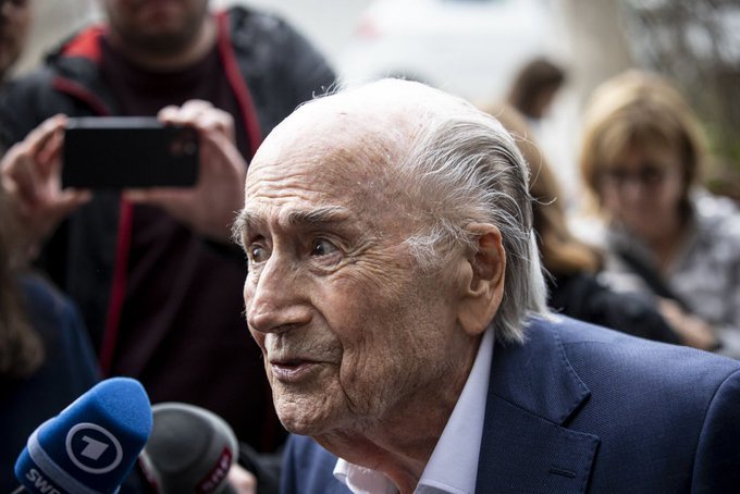 Older man with white hair in a blue suit being interviewed by reporters, microphones and a smartphone visible.
