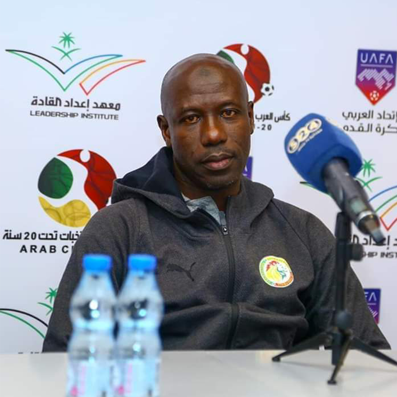 Man at a press conference wearing a dark zip hoodie, speaking into a blue microphone with logos in the background (Leadership Institute; Arab Cup). Man at a press conference wearing a dark zip hoodie, speaking into a blue microphone with logos in the background (Leadership Institute; Arab Cup).