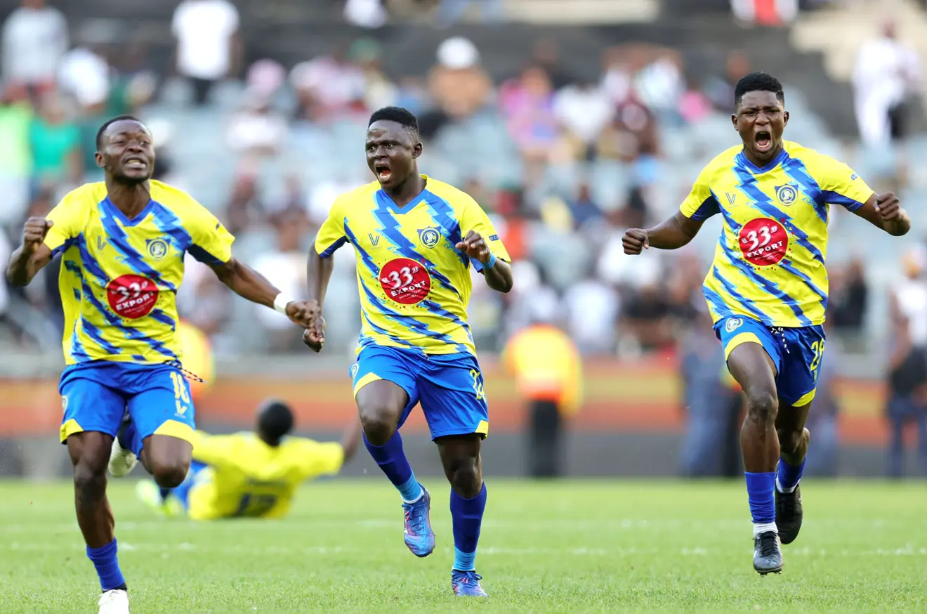 Three soccer players in yellow and blue jerseys sprint across a green field in celebration, with a blurred crowd in the stands behind them.