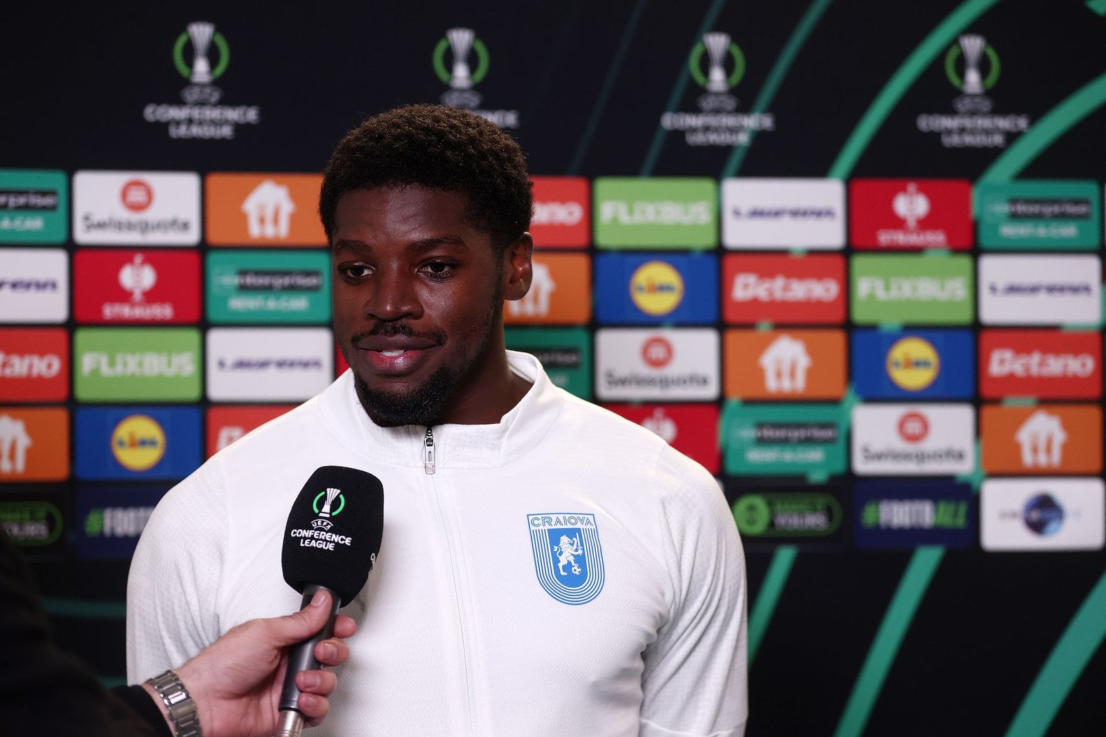 Male footballer in a white jacket being interviewed at a press conference, sponsor boards in the background. Male footballer in a white jacket being interviewed at a press conference, sponsor boards in the background.