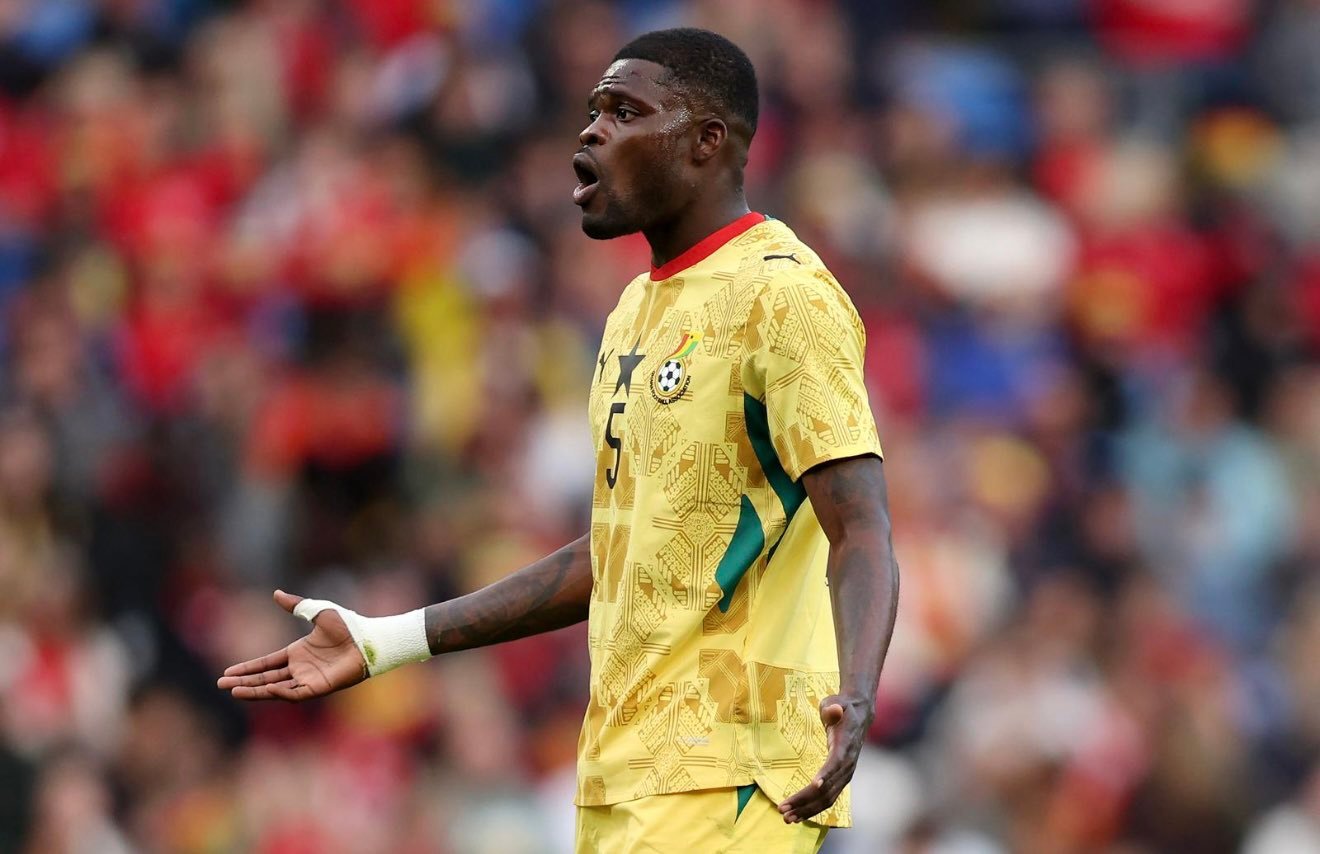 Ghanaian footballer in a yellow kit with number 5 on the chest, arms outstretched on the field during a match, stadium crowd in the background.