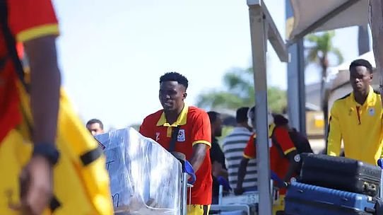 Athlete in red jersey pushing a large wrapped crate on a wheeled cart at an outdoor event, with teammates nearby. Athlete in red jersey pushing a large wrapped crate on a wheeled cart at an outdoor event, with teammates nearby.