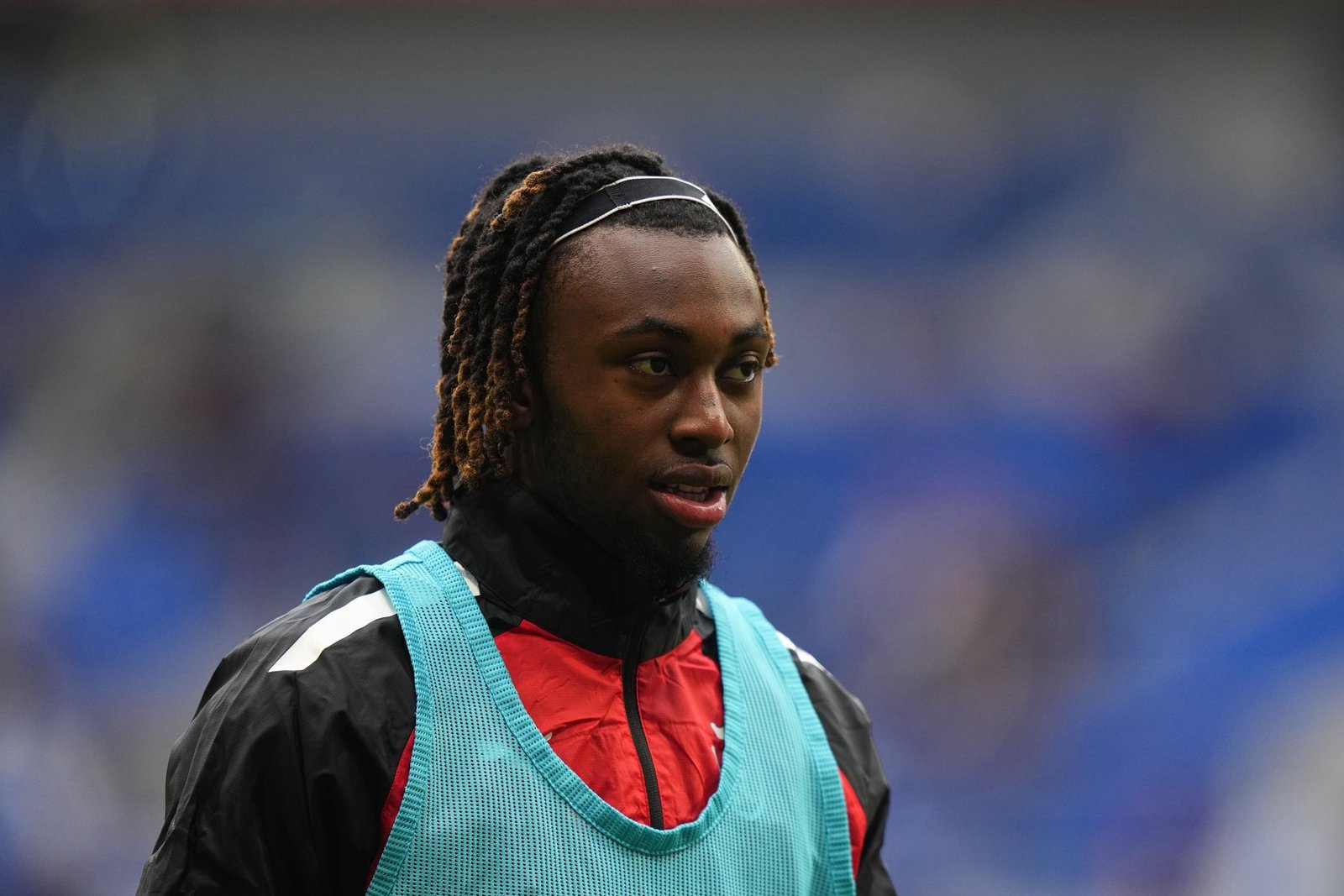 Athlete with braided hair wearing a turquoise training bib over a red and black jacket, looking off to the side.