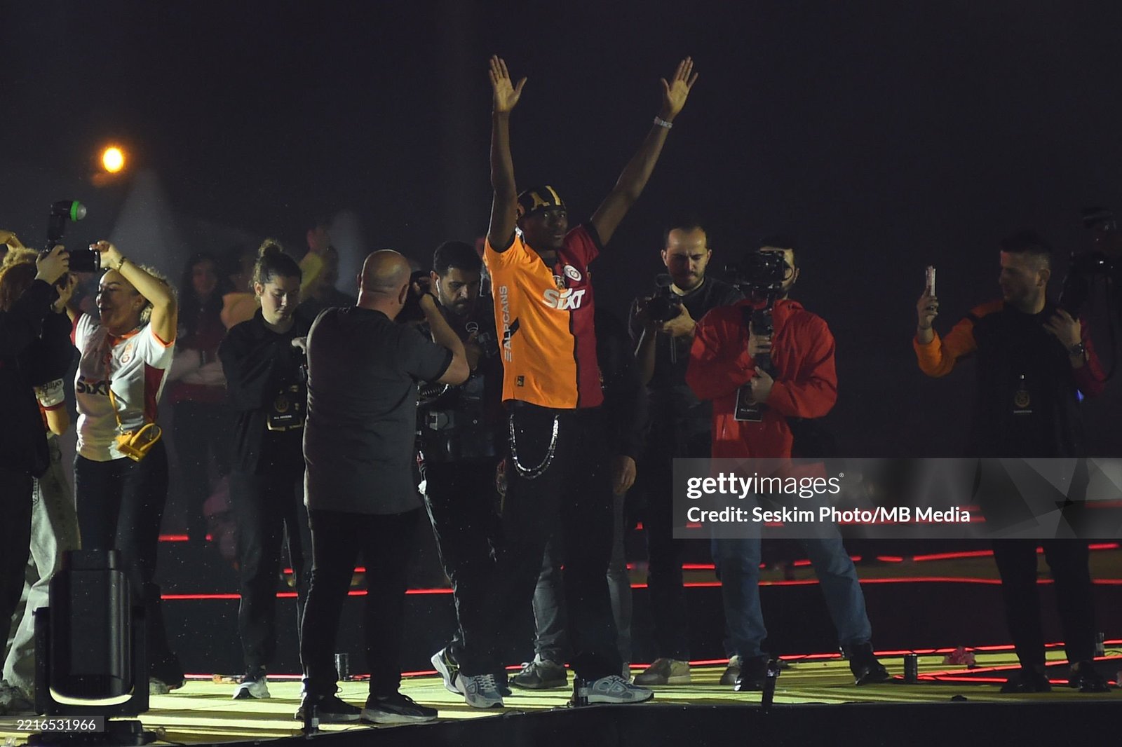 Athlete in orange and red raises arms in victory on a stage while photographers capture the moment at night. Athlete in orange and red raises arms in victory on a stage while photographers capture the moment at night.