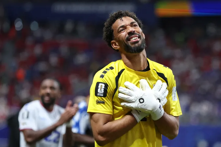 Soccer goalkeeper in a bright yellow kit holds his chest with gloved hands, smiling on the field.