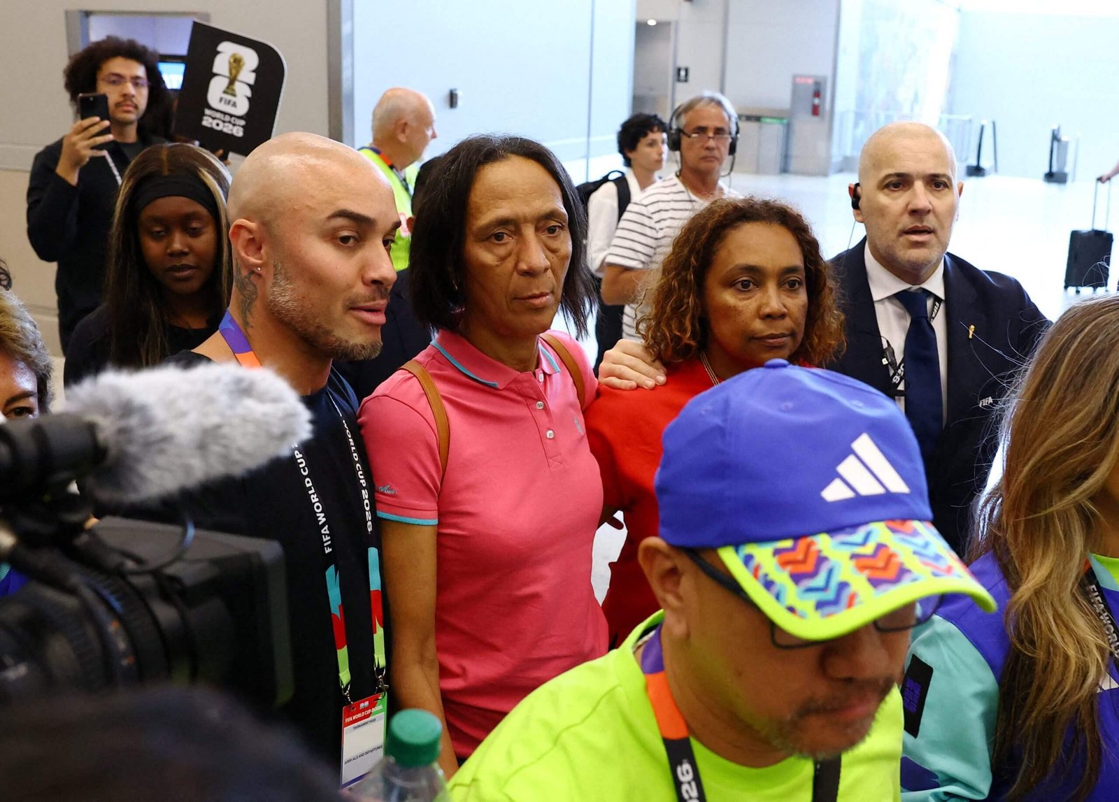 Group of diverse people in a bright indoor lobby crowds around a central man in a pink polo, wearing lanyards and tags for an event.