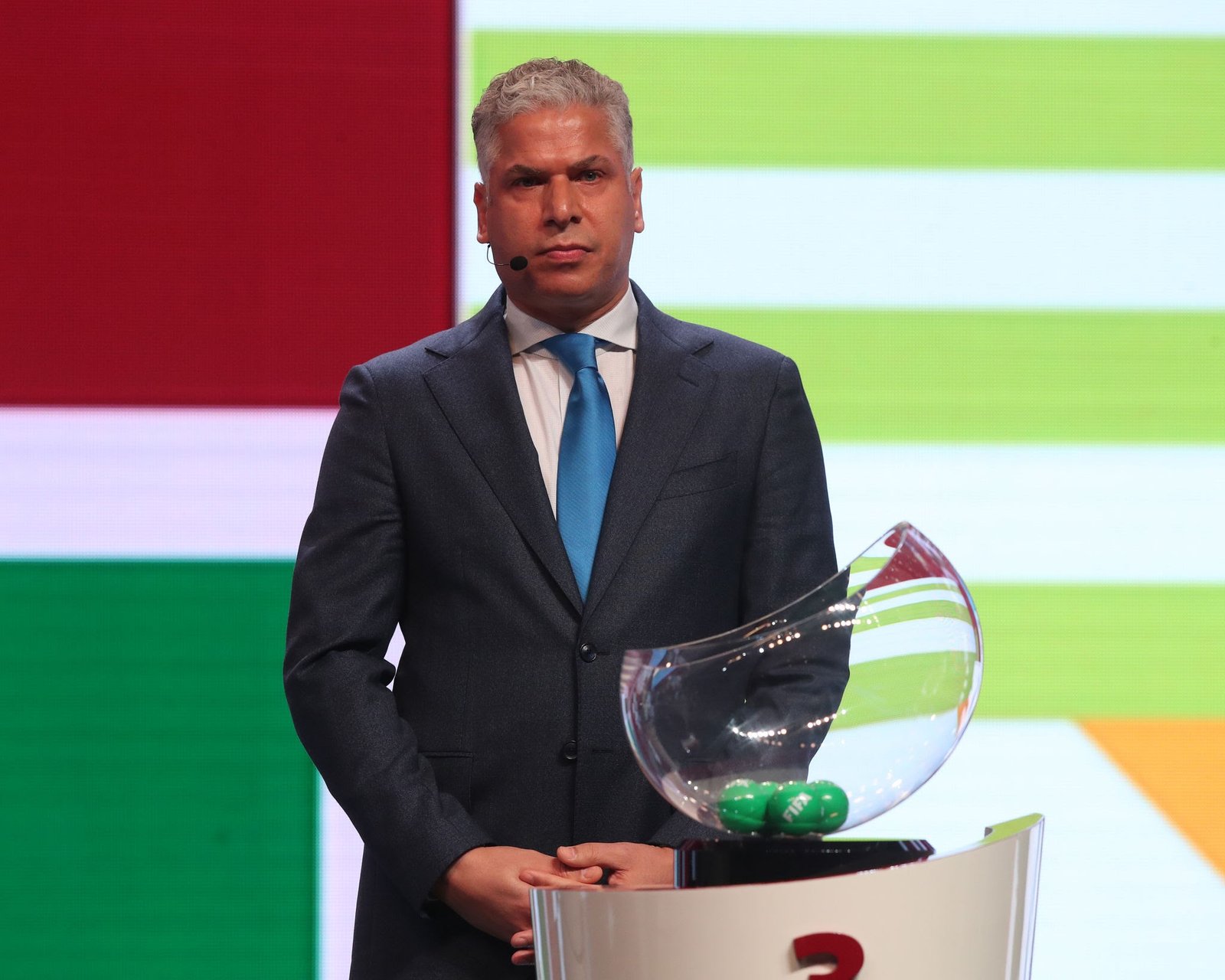 Man in a dark suit with blue tie standing at a podium, wearing a headset microphone, against a bright colored backdrop holding a glass award. Man in a dark suit with blue tie standing at a podium, wearing a headset microphone, against a bright colored backdrop holding a glass award.