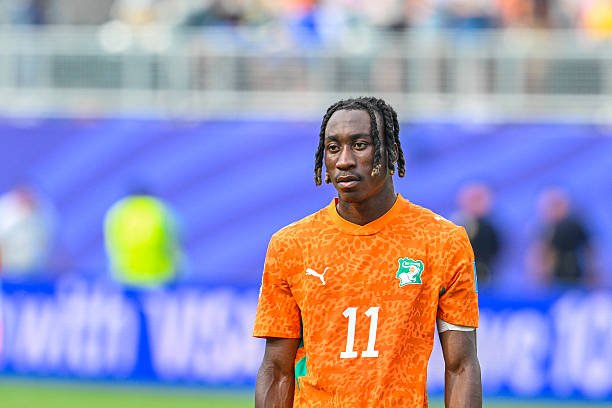 Soccer player wearing an orange jersey with the number 11 stands on a grassy field during a match, with a blurred blue stadium backdrop behind him. Soccer player wearing an orange jersey with the number 11 stands on a grassy field during a match, with a blurred blue stadium backdrop behind him.