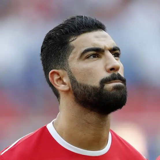 Close-up of a male soccer player with dark hair and a full beard, wearing a red jersey, gazing upward. Close-up of a male soccer player with dark hair and a full beard, wearing a red jersey, gazing upward.