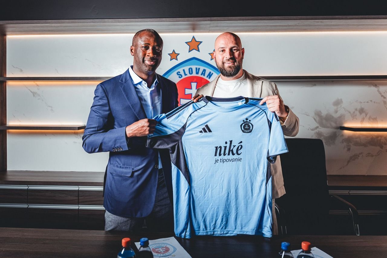 Two men in suits present a light-blue football jersey with Nike logo and a crest in a conference room.