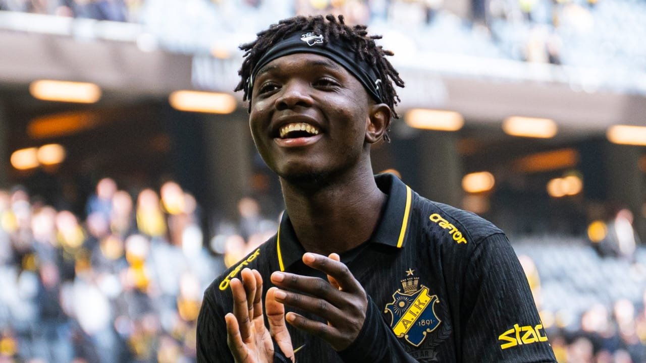 Young footballer in a black jersey claps and smiles on the field, stadium lights in the background.
