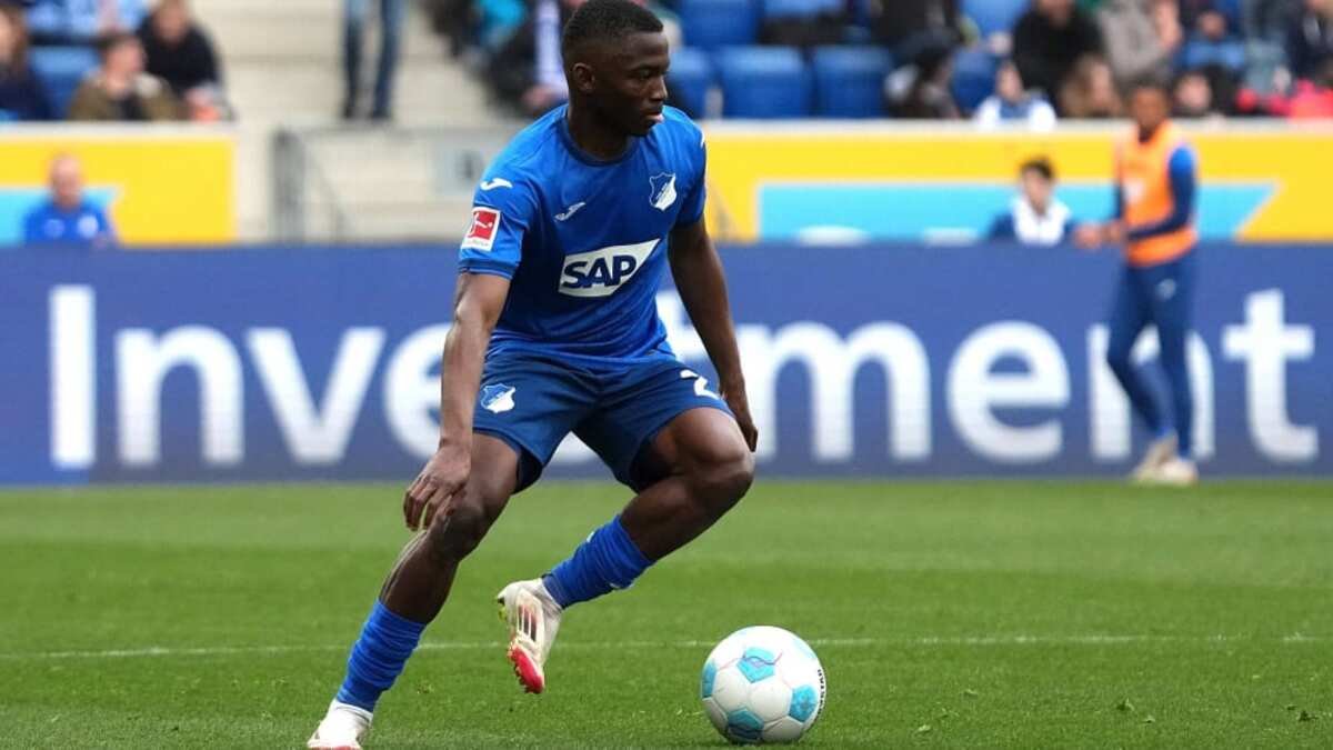 Soccer player in a blue uniform dribbling a white and blue ball on a green pitch with stadium signage in the background.