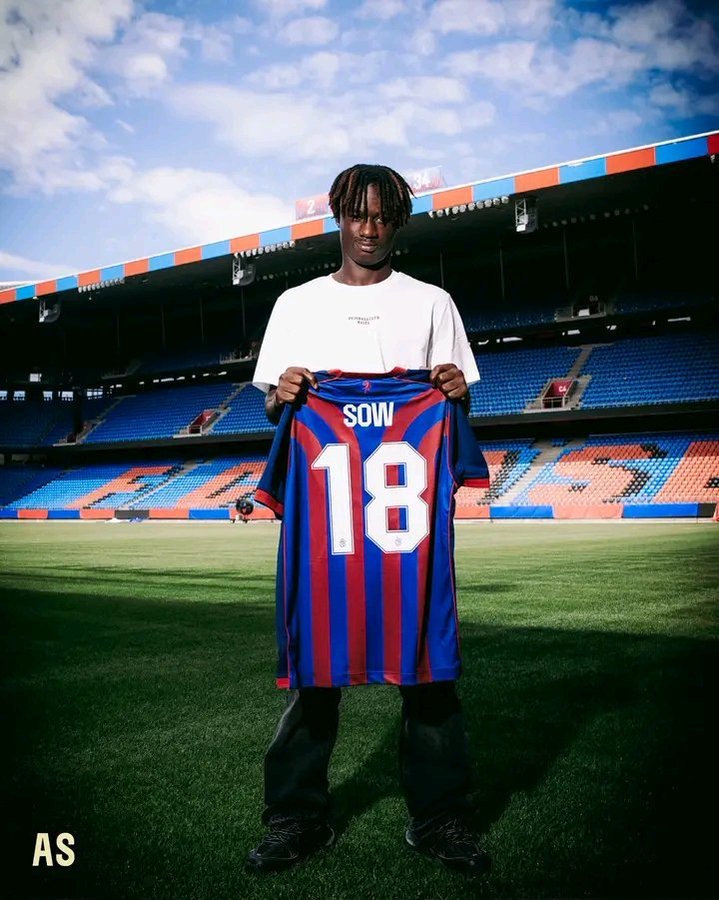 Young man on a football pitch holding a blue-and-red striped jersey with the name 'SOW' and number 18 in front of empty stadium seating.