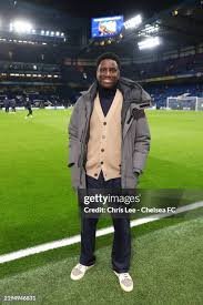 Man stands on a soccer field inside a large stadium at night, wearing a beige cardigan under a gray puffer jacket, dark pants, and light sneakers, with bright lights and seating visible in the background.