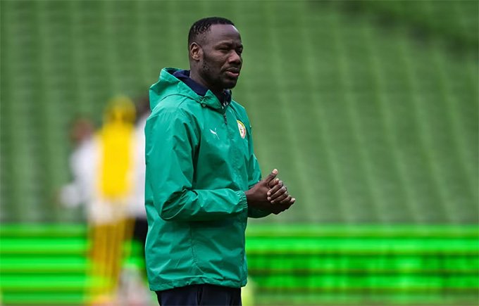 Sports coach in a green jacket standing on a football training field with blurred stands in the background, hands clasped together. Sports coach in a green jacket standing on a football training field with blurred stands in the background, hands clasped together.