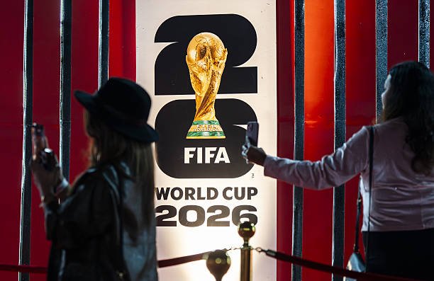 Two people pose for selfies beside a FIFA World Cup 2026 banner featuring a gold trophy against a red backdrop. Two people pose for selfies beside a FIFA World Cup 2026 banner featuring a gold trophy against a red backdrop.