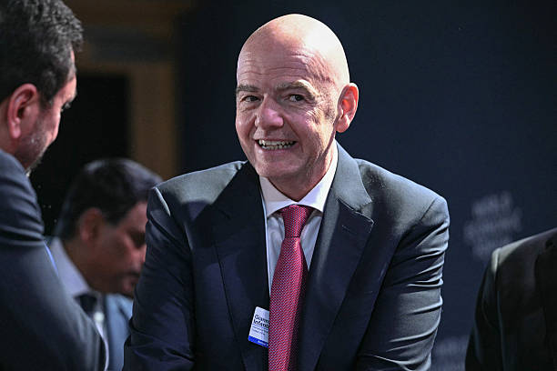 Bald man in a dark suit and pink tie smiles while conversing at a formal event, with others in the background. Bald man in a dark suit and pink tie smiles while conversing at a formal event, with others in the background.