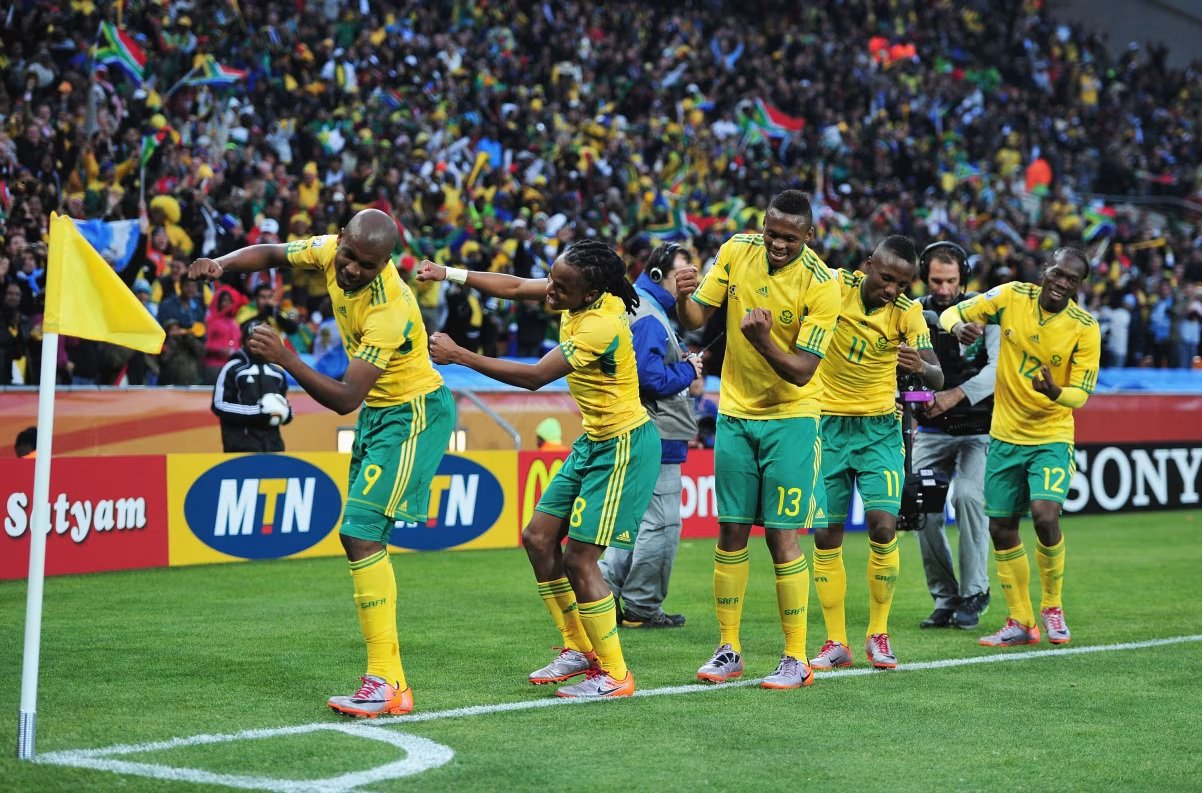 South Africa national football team players in yellow and green celebrate near the corner flag as a packed stadium cheers.