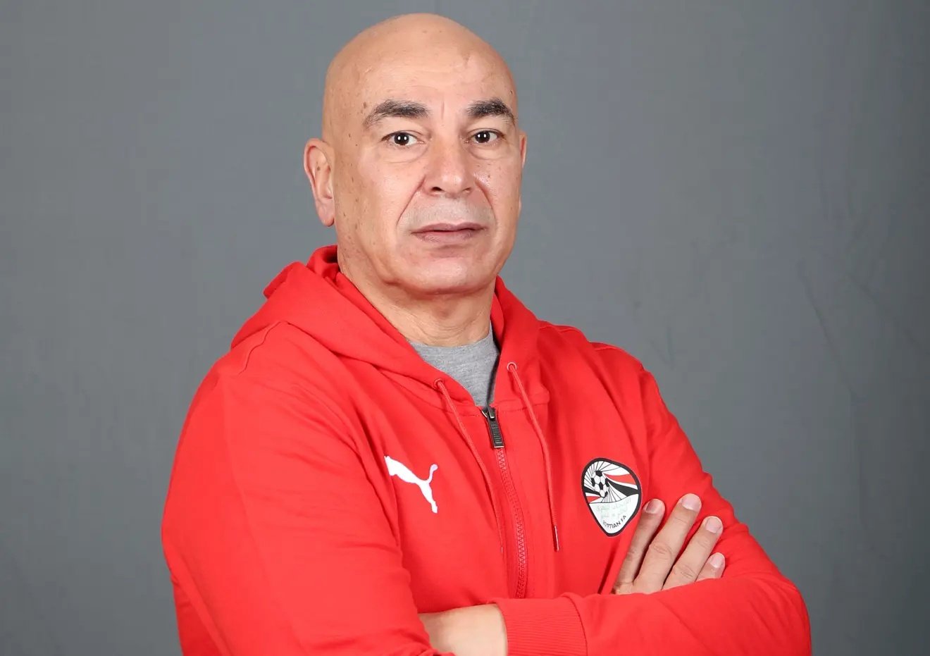 Portrait of a middle-aged man with a shaved head, wearing a red hoodie, arms crossed, against a neutral gray background.
