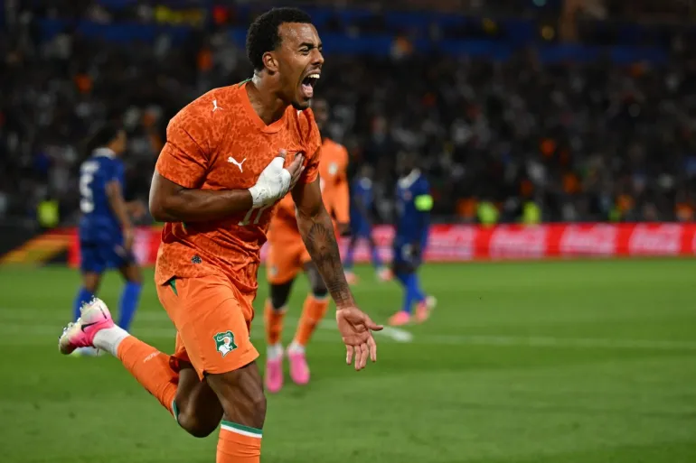 Soccer goalkeeper in an orange kit celebrates on the field with a fist pump.