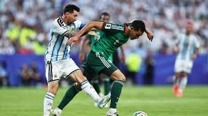 Two soccer players chase the ball during a match; one wears a light blue-and-white striped jersey and the other a green jersey, with a crowd in the background.