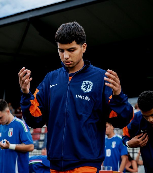 Soccer player in a blue Netherlands training jacket with crest, hands raised and focused stance.