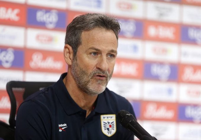 Male sports official with gray beard speaks at a press conference, microphone in front and sponsor backdrop behind him.