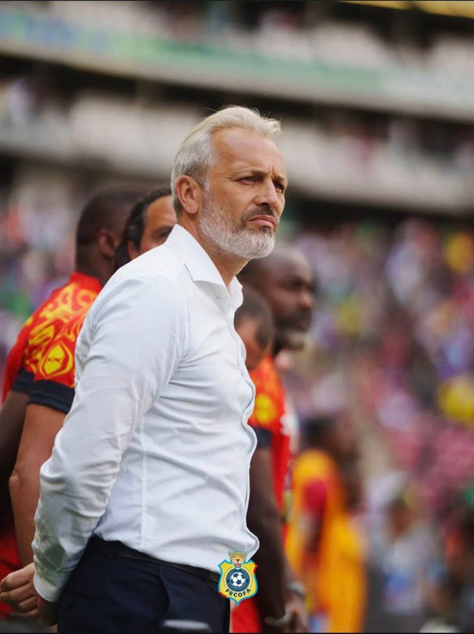 Soccer coach with gray beard in a white shirt on the sideline, focused on the game as players stand behind him.