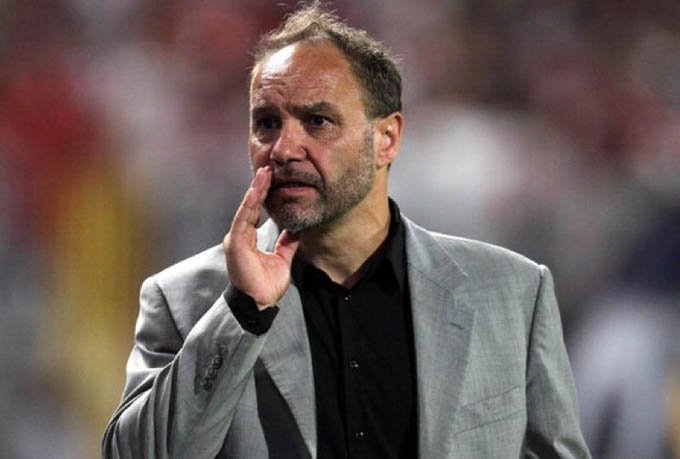 Man in a grey blazer and black shirt speaks on the field, hand raised to his mouth as if signaling. Man in a grey blazer and black shirt speaks on the field, hand raised to his mouth as if signaling.