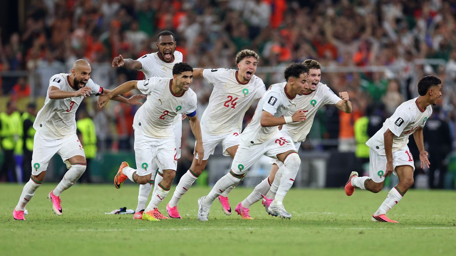 Morocco national soccer team celebrating a goal on the field, players sprinting with fans cheering in the stands behind them