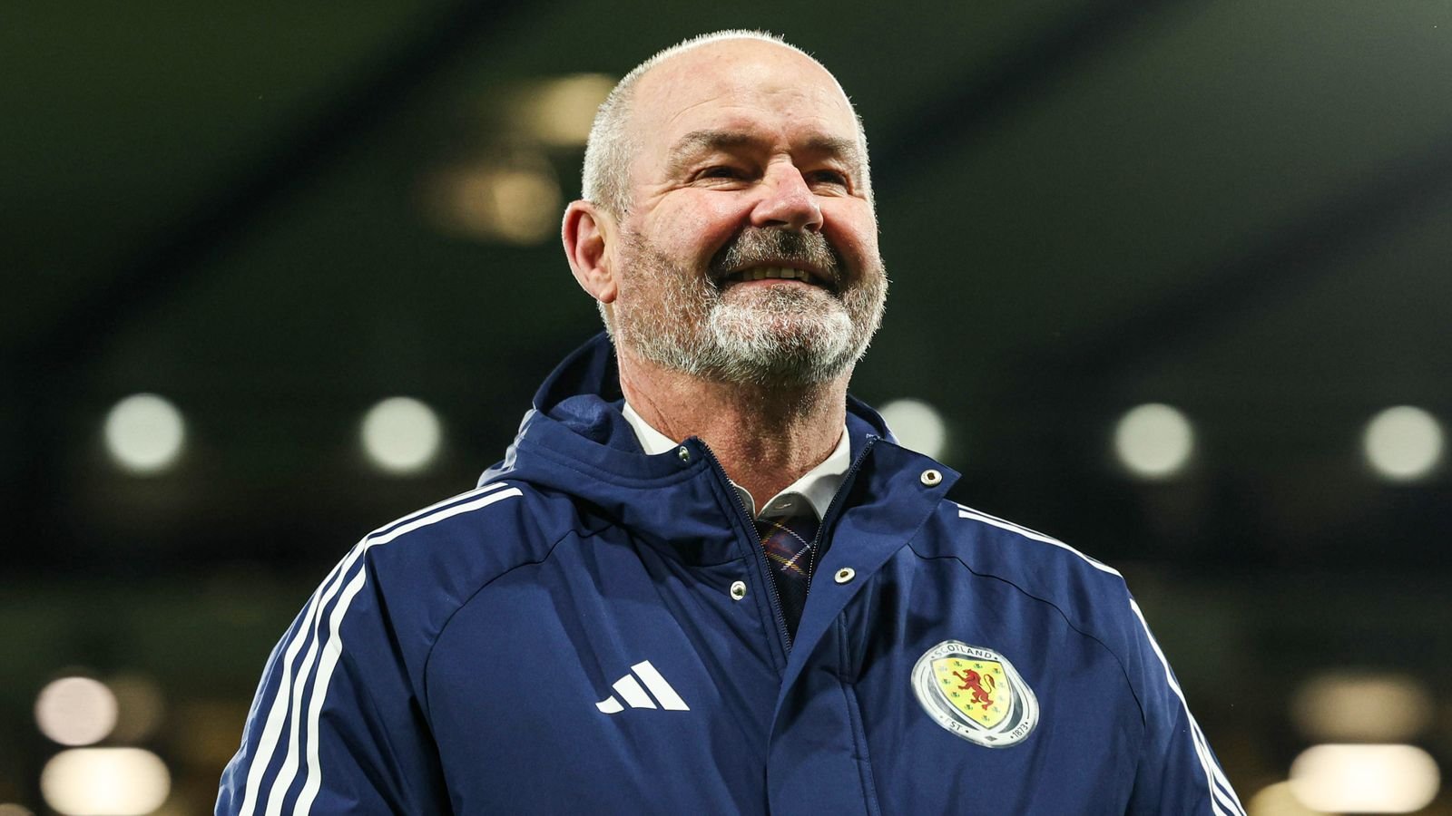 Smiling man in a Scotland football jacket with crest, outdoors at night.