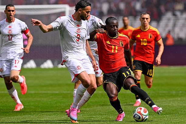 Two soccer players battle for the ball midfield during a match, one in white with number 9 and the other in red with number 11, on a green pitch.