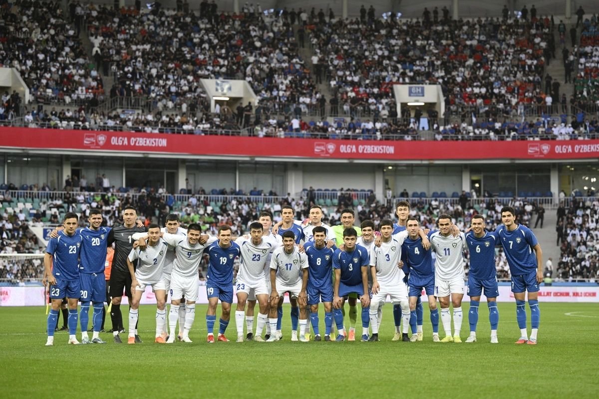 Soccer team posing for a group photo on the field with players in white and blue kits, stadium stands full of spectators behind them.