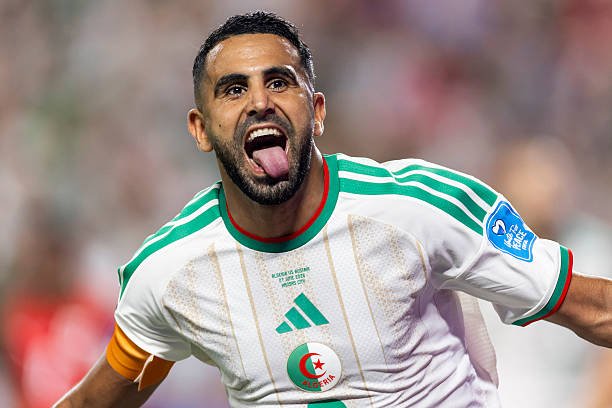 Algerian footballer celebrating a goal, tongue out, in a white jersey with green stripes and an orange captain's armband. Algerian footballer celebrating a goal, tongue out, in a white jersey with green stripes and an orange captain's armband.