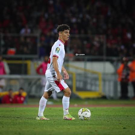 Soccer player in a white kit with red accents controls the ball on a grassy field during a match, stadium in the background. Soccer player in a white kit with red accents controls the ball on a grassy field during a match, stadium in the background.
