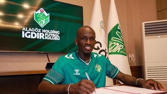 Footballer signs a contract at a club table, with the team's green jersey and club logo on a screen behind him. Footballer signs a contract at a club table, with the team's green jersey and club logo on a screen behind him.