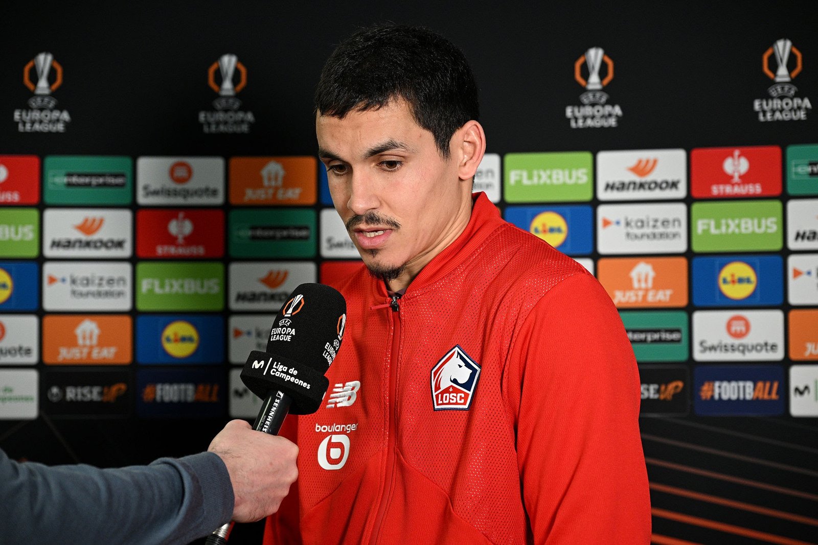 Male soccer player in a red jersey being interviewed while a sponsor backdrop with logos is visible behind him into a microphone appears before him. Male soccer player in a red jersey being interviewed while a sponsor backdrop with logos is visible behind him into a microphone appears before him.