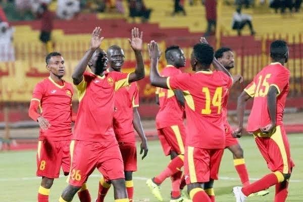 Soccer players in red and yellow jerseys celebrate on the field, raising hands in a group high-five amid a stadium backdrop.