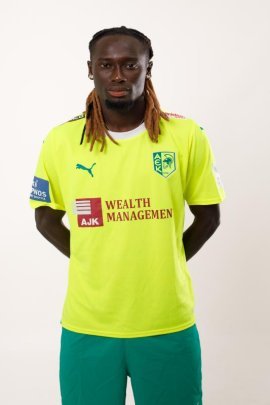 Portrait of a male footballer standing with hands behind his back, wearing a neon yellow jersey with sponsor logos and teal shorts. Portrait of a male footballer standing with hands behind his back, wearing a neon yellow jersey with sponsor logos and teal shorts.