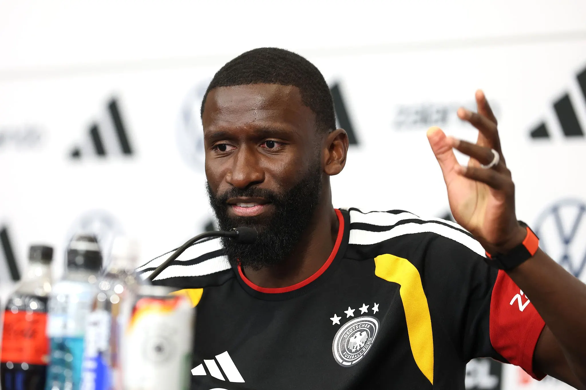 Bearded man in a German national team jersey speaks at a press conference, gesturing with his right hand. Bearded man in a German national team jersey speaks at a press conference, gesturing with his right hand.