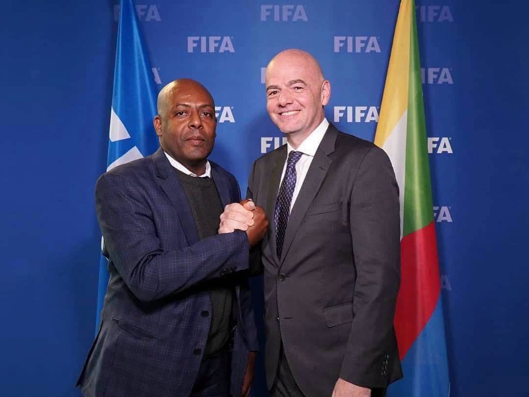Two men in business suits shake hands in front of a blue FIFA backdrop with flags on either side.