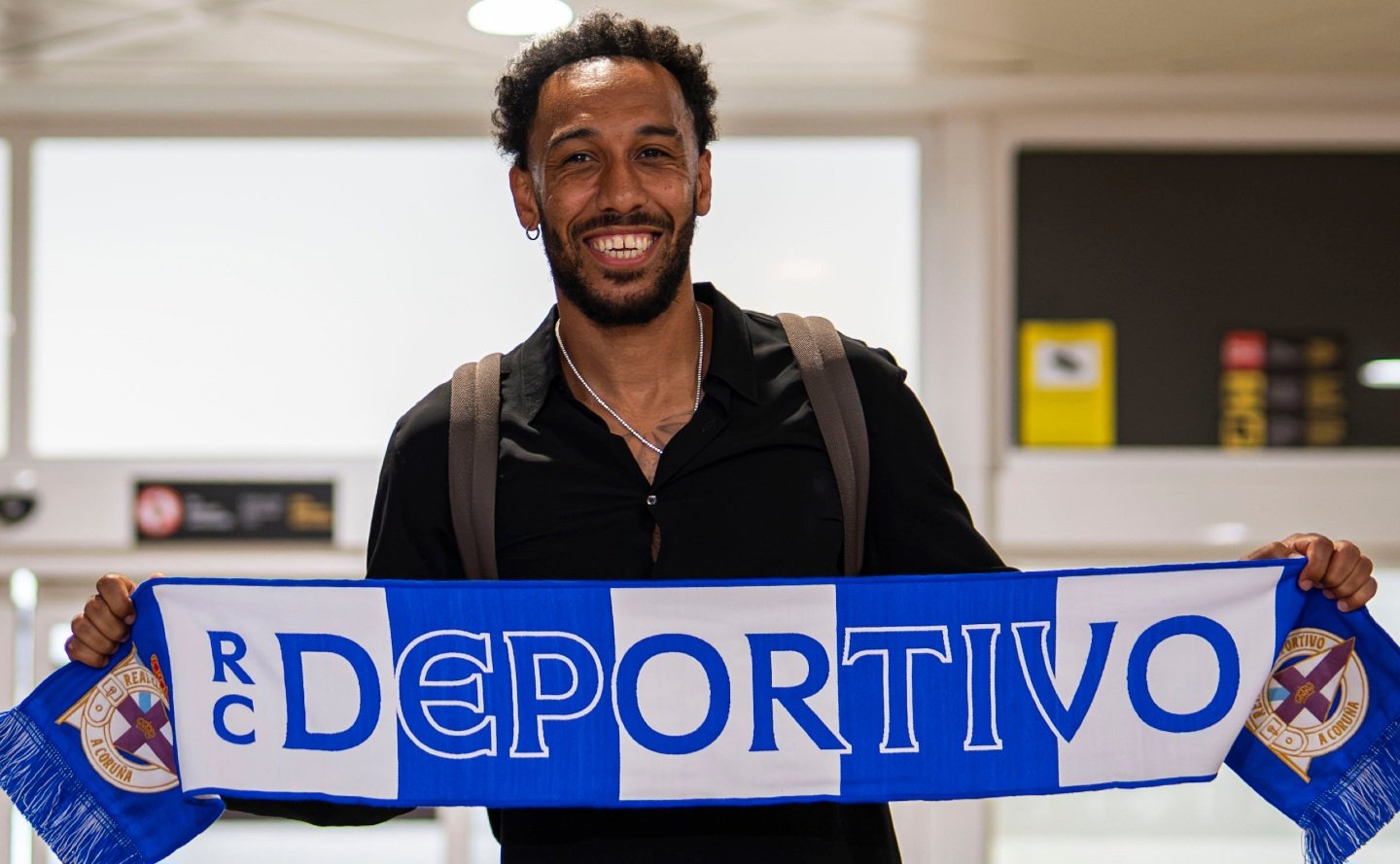 Smiling man in a black shirt holding a blue-and-white RC Deportivo scarf across his chest inside a building. Smiling man in a black shirt holding a blue-and-white RC Deportivo scarf across his chest inside a building.