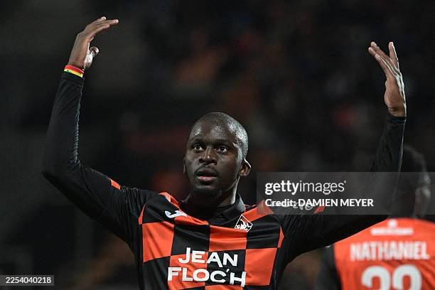 Soccer player in a black-and-orange kit raises both arms in celebration on the pitch, with a blurred crowd behind him. Soccer player in a black-and-orange kit raises both arms in celebration on the pitch, with a blurred crowd behind him.