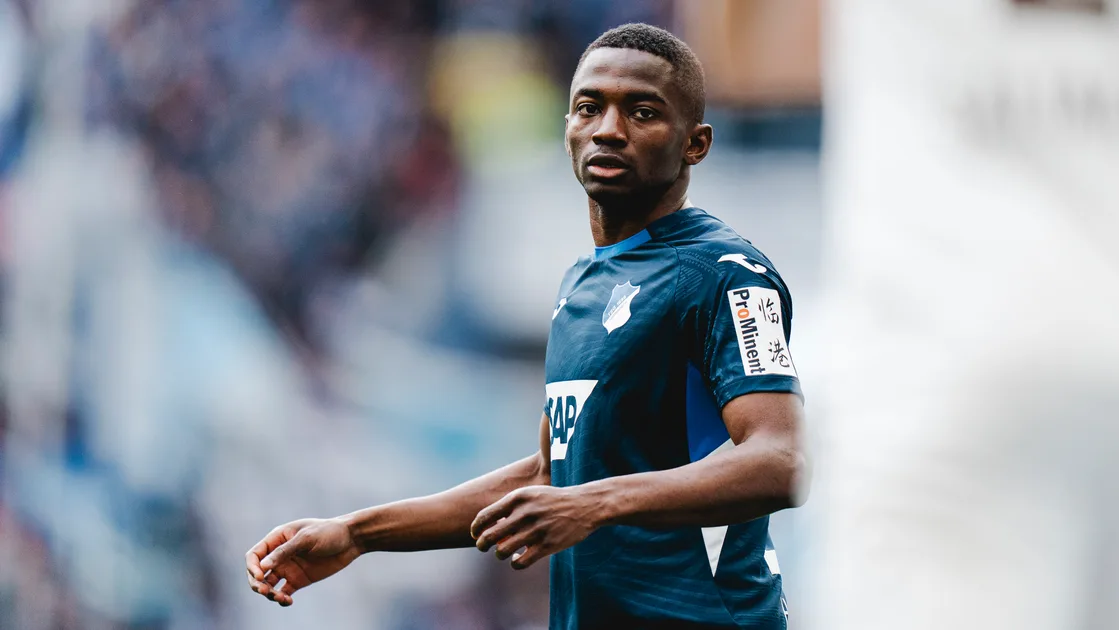 Male soccer player in a blue kit on the pitch, looking toward the camera with a focused expression. Male soccer player in a blue kit on the pitch, looking toward the camera with a focused expression.