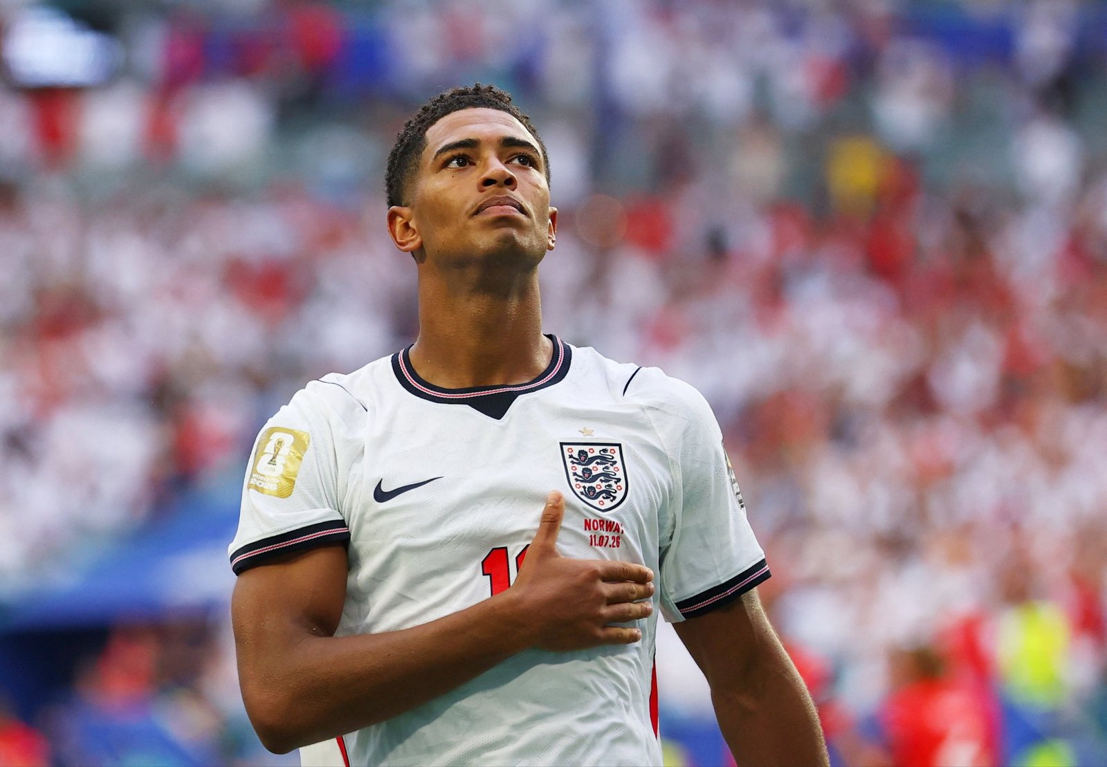 England national footballer looking up toward the sky in a white England kit, stadium crowd blurred in the background.